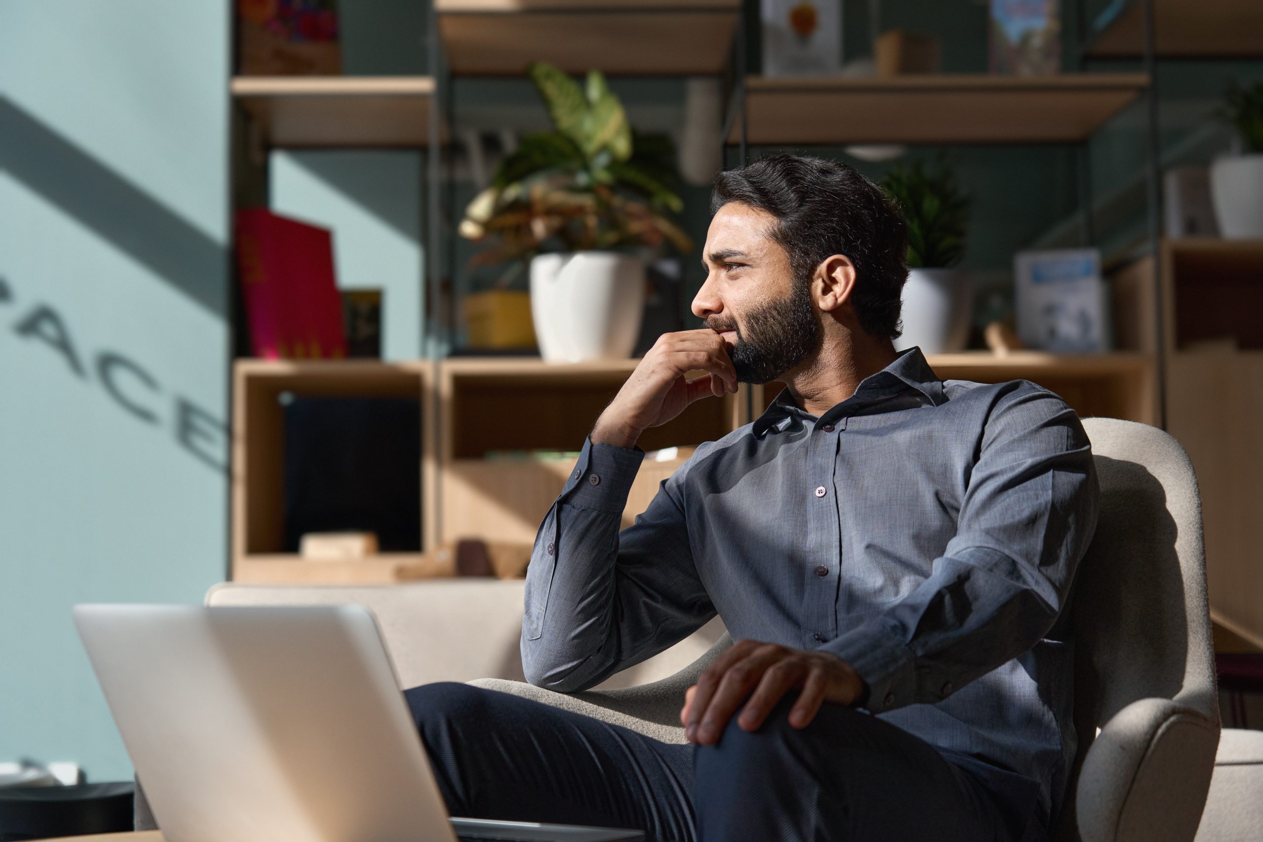 Young thoughtful smart indian leader professional business man executive looking away relaxing sitting on chair in modern office lobby with laptop, thinking of new ideas, dreaming of success, planning.